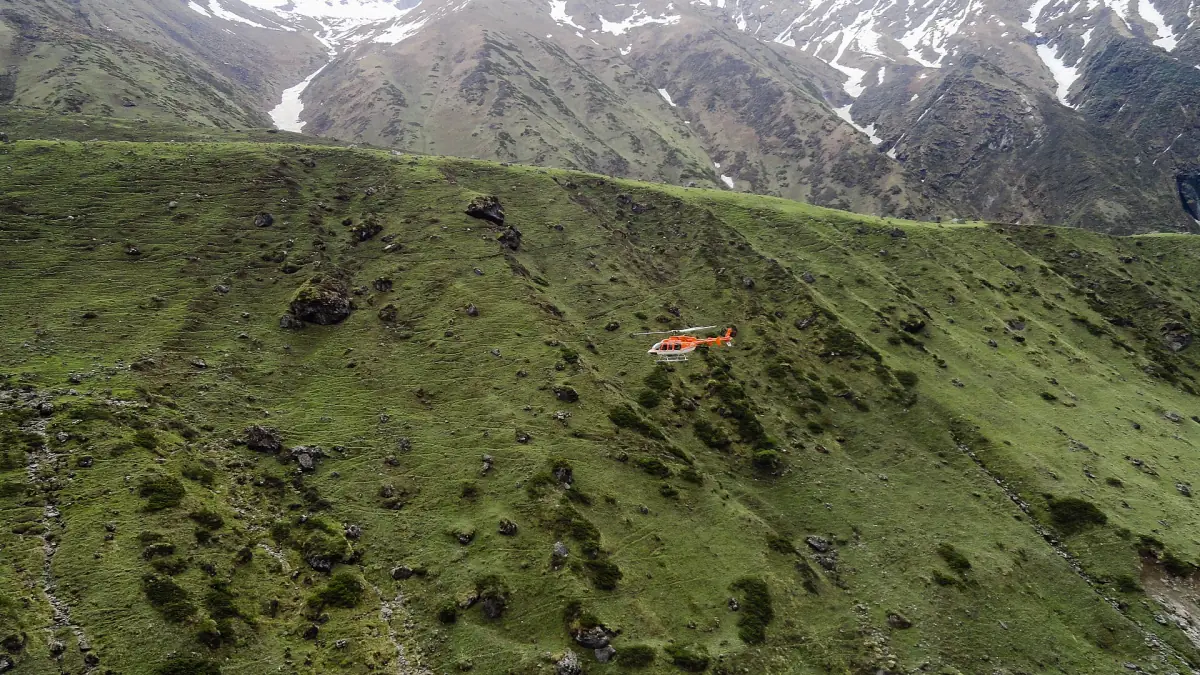 Small helicopter flying over a green mountain slope with snow peaks behind
